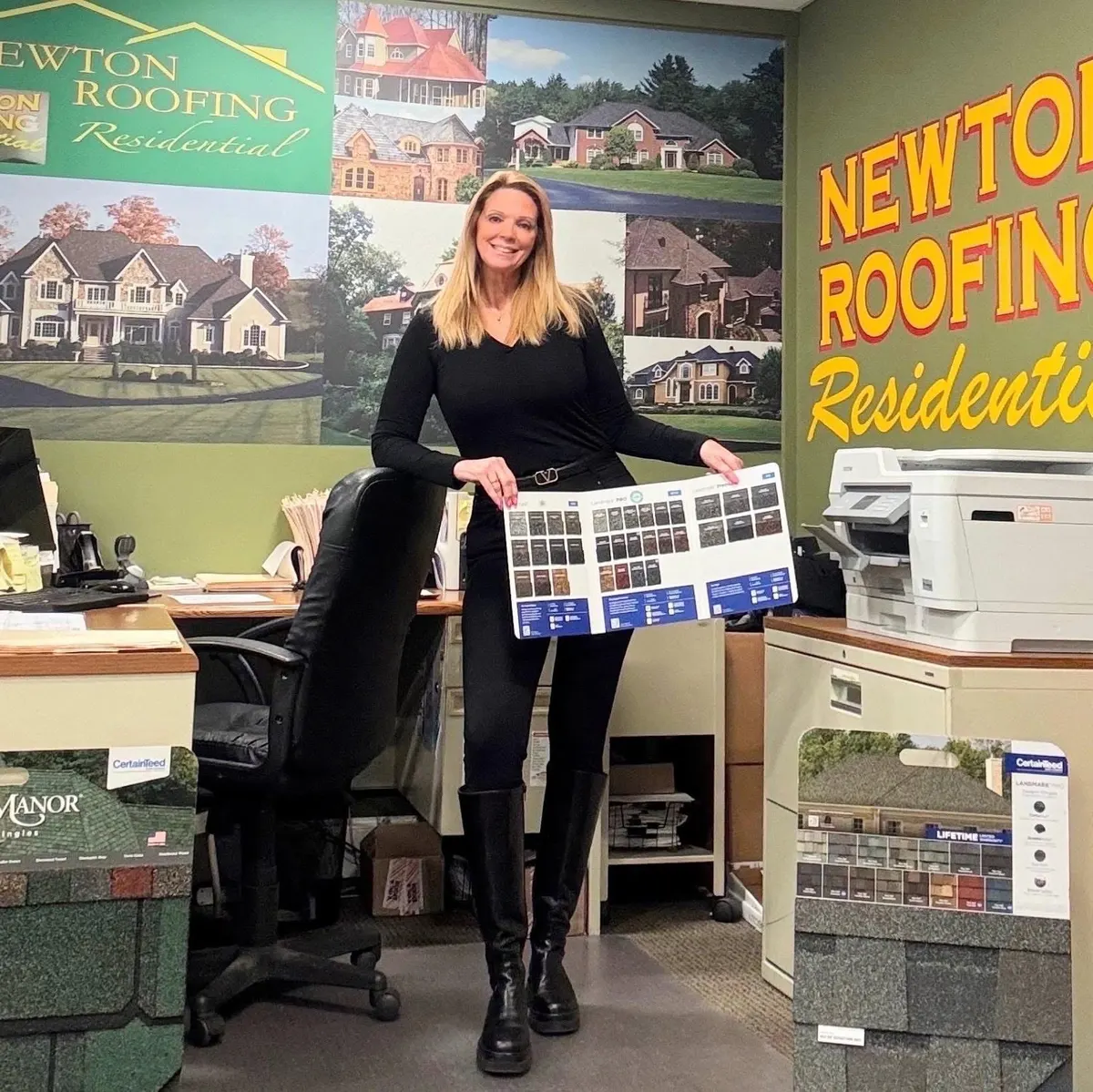 Toni Bryan in the Newton Roofing office holding a shingle color selector board, beside displays of CertainTeed shingle samples